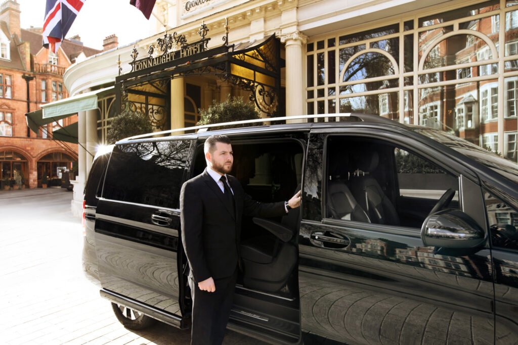 A professional chauffeur in a black suit holds open the sliding door of a black Mercedes-Benz V-Class van in front of the prestigious Connaught Hotel.