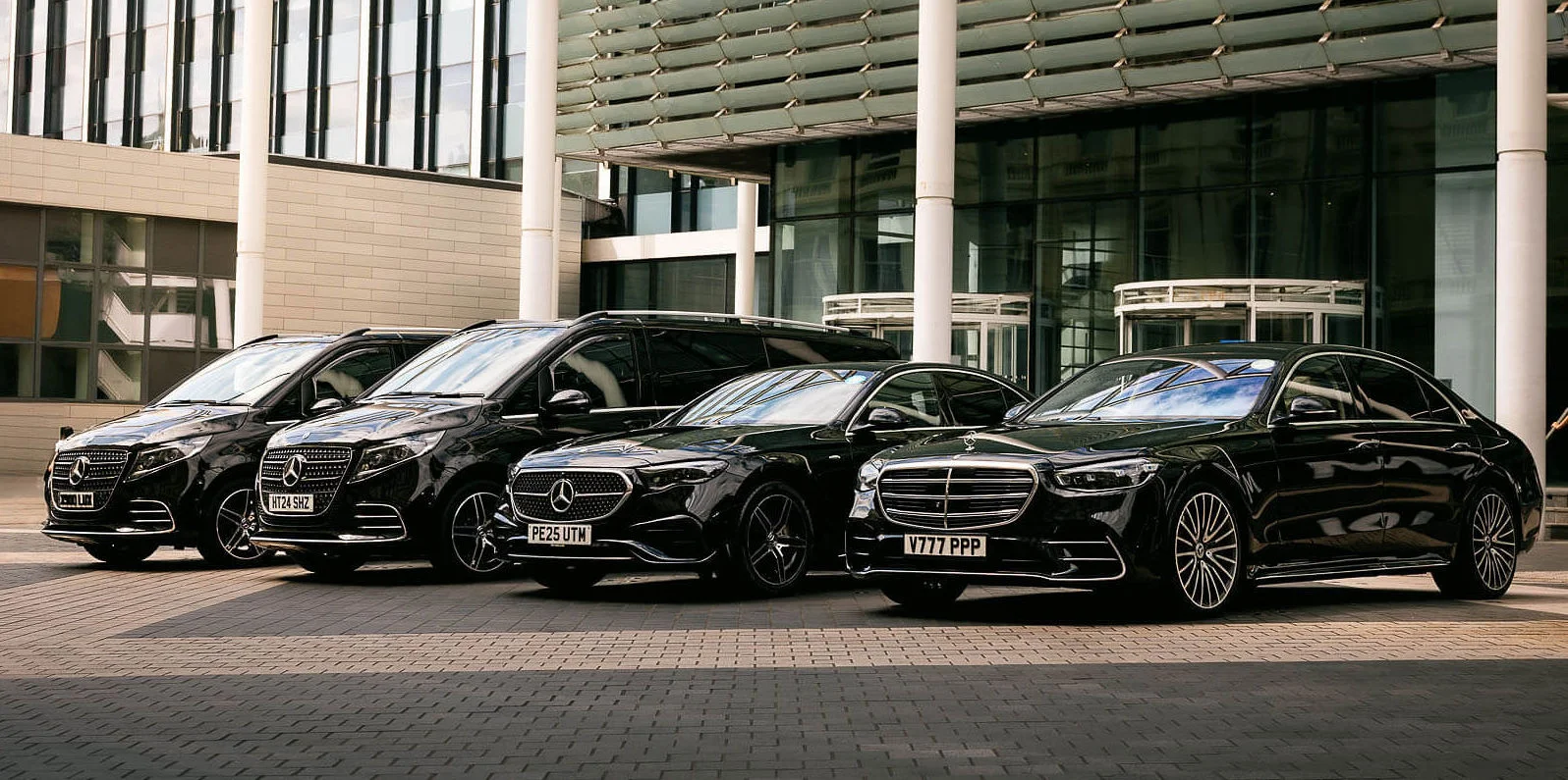 A lineup of four black Mercedes-Benz luxury vehicles, including two V-Class vans and two premium sedans, parked in front of a modern glass building.